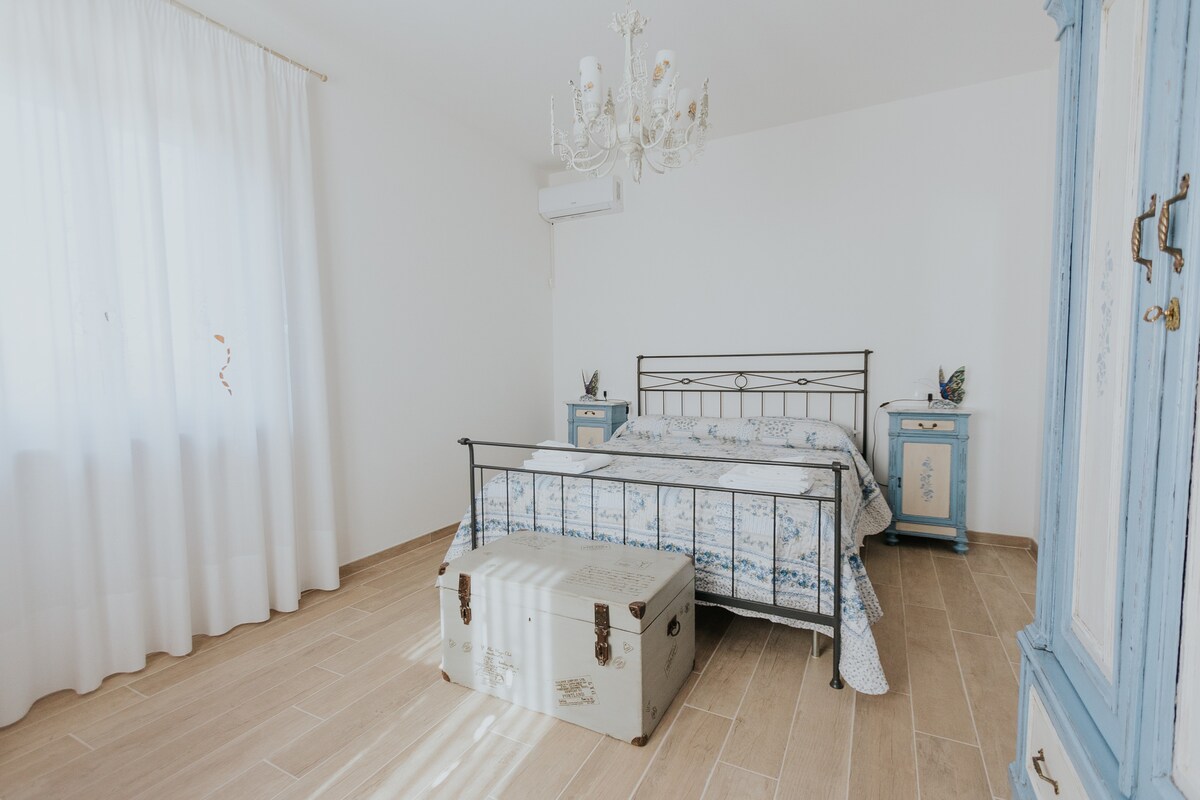 A bright bedroom is presented with a large window draped in sheer white curtains, allowing natural light to fill the space. A metal bed frame is complemented by a floral bedspread. Vintage nightstands with decorative details and a trunk are positioned at the foot of the bed.
