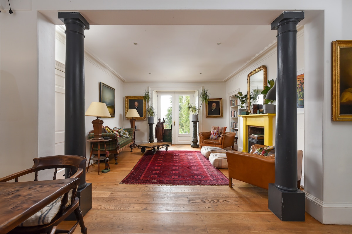 An elegant living area showcases a rich area rug on hardwood flooring, framed by dark columns. A mix of comfortable seating, including a leather sofa and armchairs, is arranged around a central fireplace, while natural light filters through large windows, illuminating the artwork and plants throughout the space.