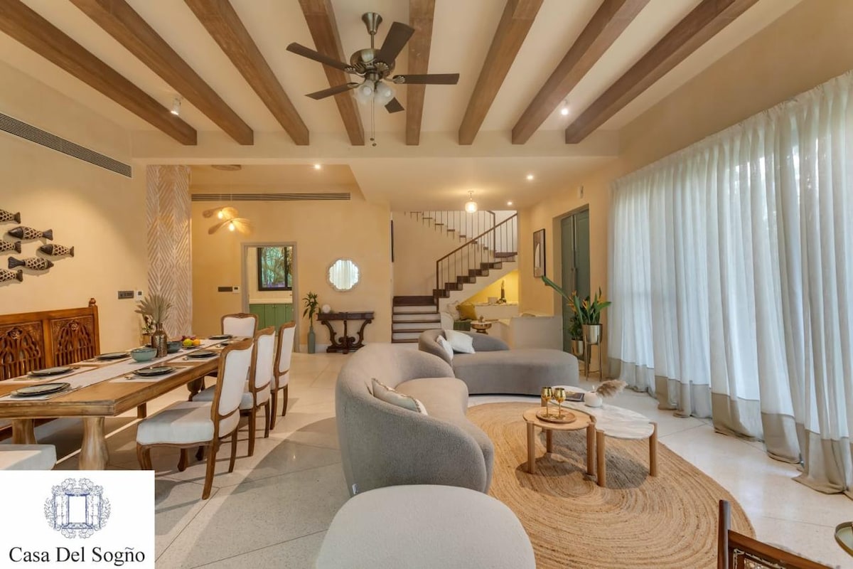 An open living space features a blend of modern and traditional furnishings, including a long dining table and curved sofas. Natural light filters in through expansive windows, while wooden beams and a ceiling fan add to the airy feel. A staircase leads to the upper level.