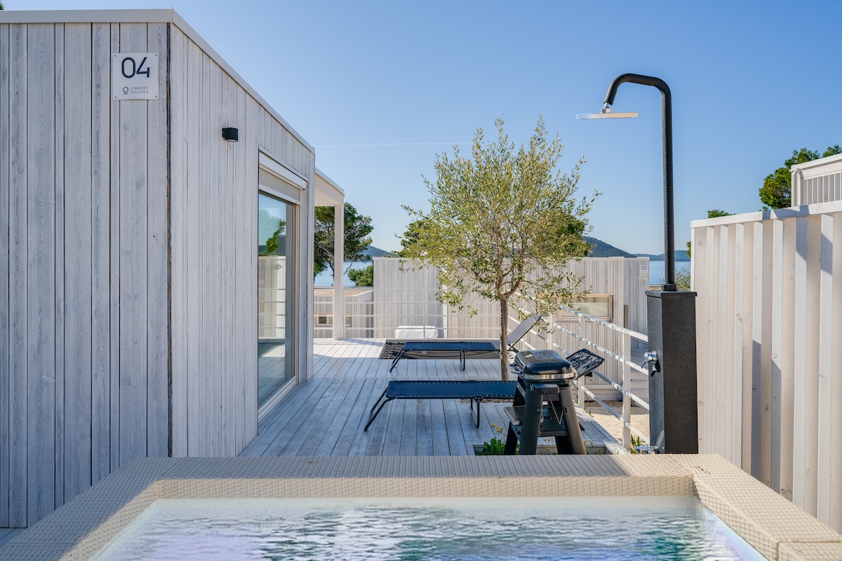 An outdoor area features a hot tub surrounded by wooden decking. Sun loungers are positioned nearby, accompanied by a sleek outdoor shower. A tree adds a touch of greenery, while a barbecue grill is available for outdoor cooking, all under a clear blue sky.