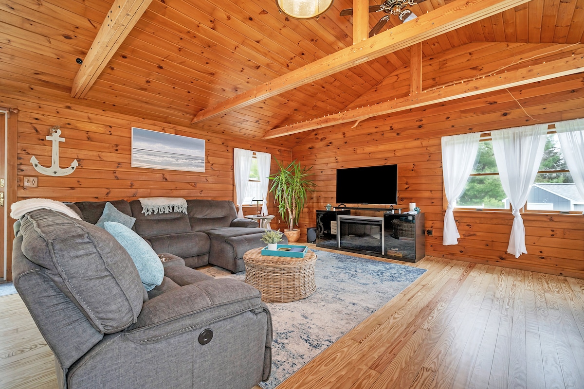 A cozy living area is surrounded by warm wooden walls, featuring a large sectional sofa and a circular woven coffee table. Light streams through sheer white curtains. A flat-screen TV is mounted on the wall, complemented by a tall indoor plant and nautical-themed decor.