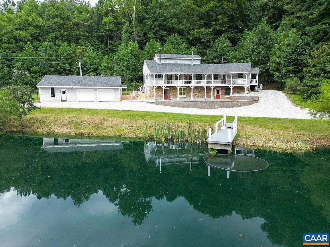 The exterior of a handbuilt unit is visible, situated beside a tranquil pond. A gravel driveway leads to the main entrance, while the balcony extends along the second floor. Surrounded by lush greenery, the setting offers a peaceful retreat in nature.
