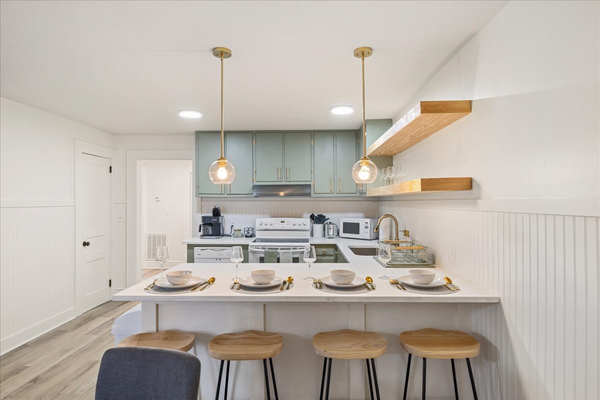 The kitchen is fully equipped with modern appliances and features a spacious countertop. Four wooden stools are arranged at the breakfast bar. Pendant lights hang above, illuminating the area. Shelves hold glassware, enhancing the kitchen's functionality.