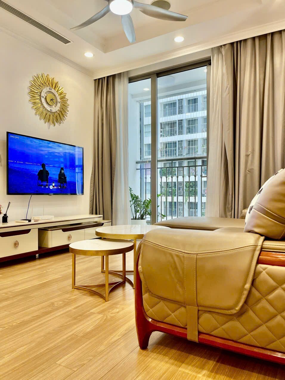 A stylish living room features a comfortable sofa and a wall-mounted television displaying a vibrant scene. Large windows provide natural light, complemented by sheer curtains. Two small coffee tables are placed in front of the seating area, enhancing the modern design of the space.