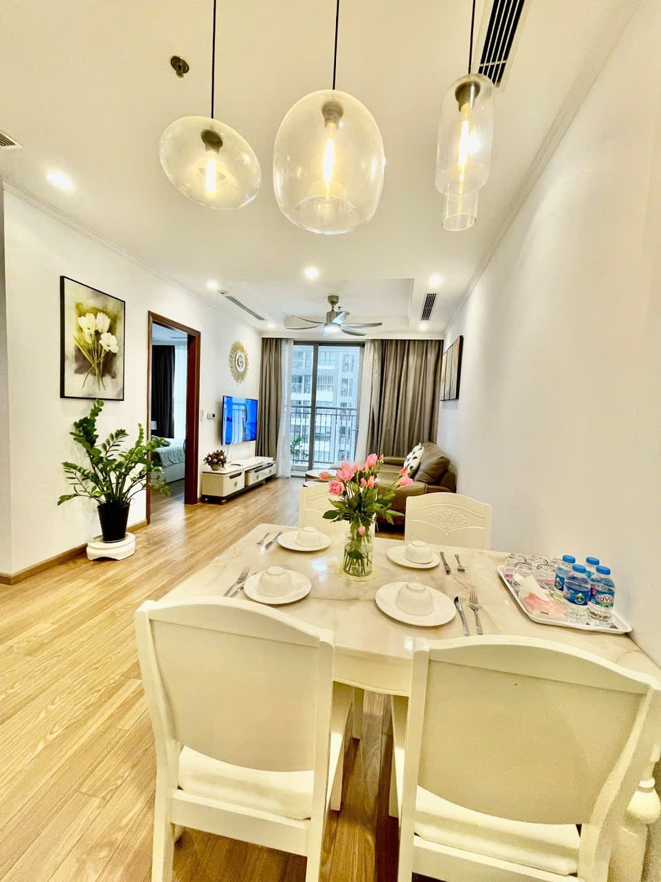 A dining area is highlighted by a marble table set for four, complete with plates and utensils. Pendant lights hang overhead, illuminating the space. A comfortable sofa and a TV are visible in the background, along with a vibrant plant adding a touch of greenery.