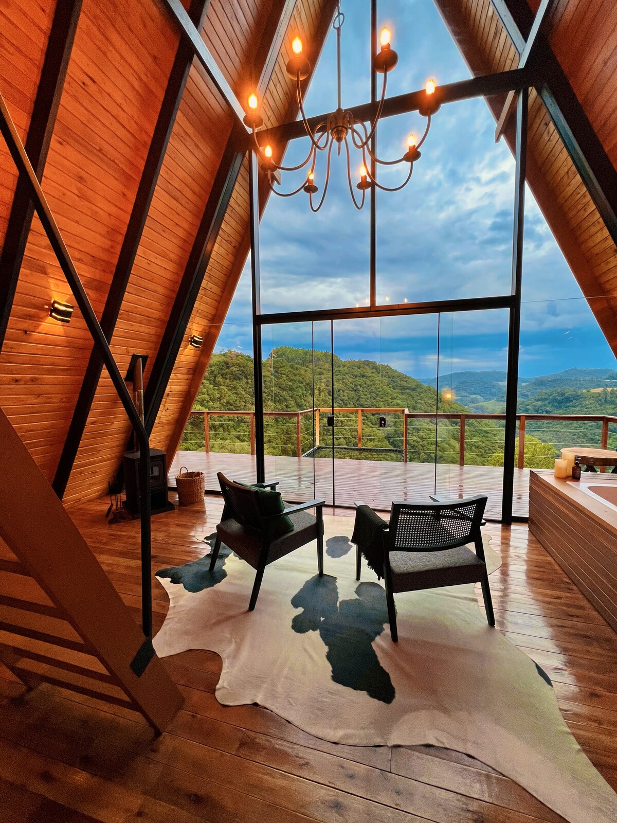A spacious living area features a striking A-frame design with large glass panels offering expansive views of the surrounding nature. Two black chairs are positioned on a cowhide rug, illuminated by a chandelier above. Natural wood accents warm the space.