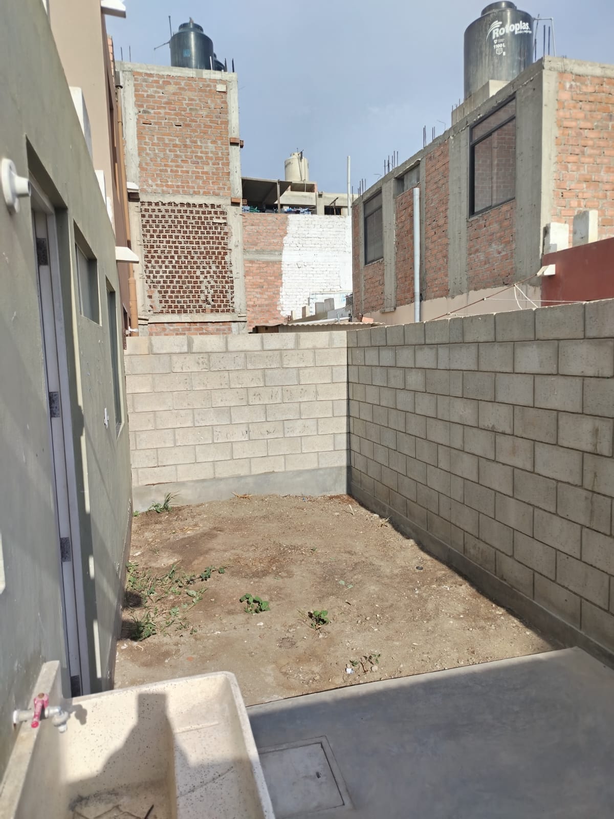A private outdoor space is shown, enclosed by gray block walls. The area is unpaved, featuring bare earth with minimal vegetation. A water faucet is visible on one wall, contributing to the functional layout of the space.