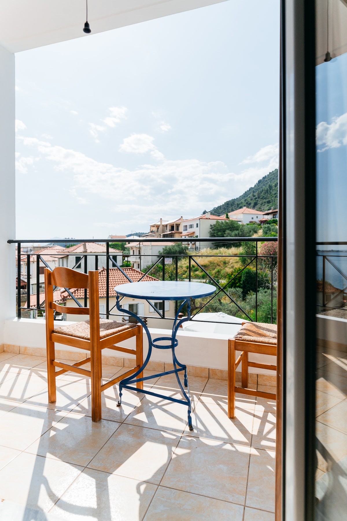 A private balcony is showcased, featuring a small table surrounded by two wooden chairs. The view includes nearby houses and lush greenery, complemented by bright sunlight and a blue sky. The railing adds a sense of openness to the outdoor space.