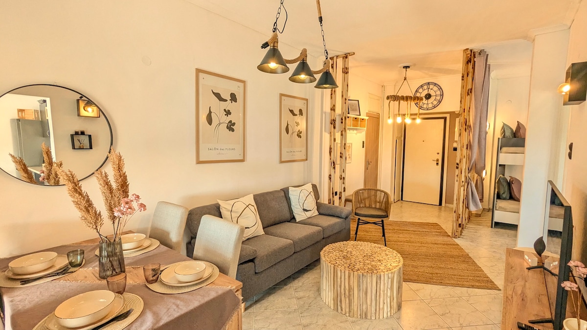 A cozy living area features a gray sofa, a circular wooden coffee table, and a meticulously set dining table. Wall art and decorative elements incorporate natural textures, while unique pendant lighting adds warmth. A neutral color palette is complemented by light-colored flooring and accents.