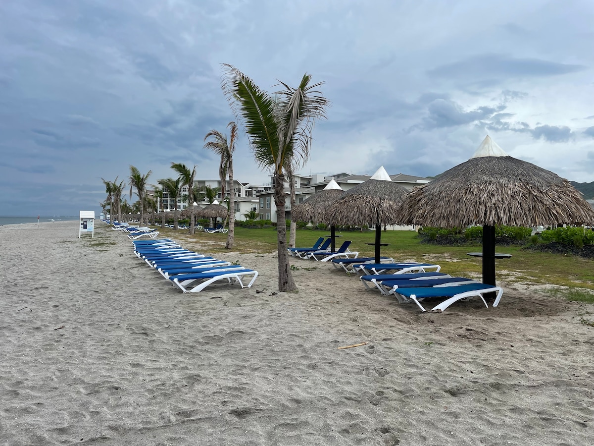 A row of blue lounge chairs is positioned on the sandy beach, shaded by thatched umbrellas. Palm trees gently sway in the breeze, with a backdrop of a resort building visible in the distance. Grey clouds fill the sky, adding to the serene coastline atmosphere.