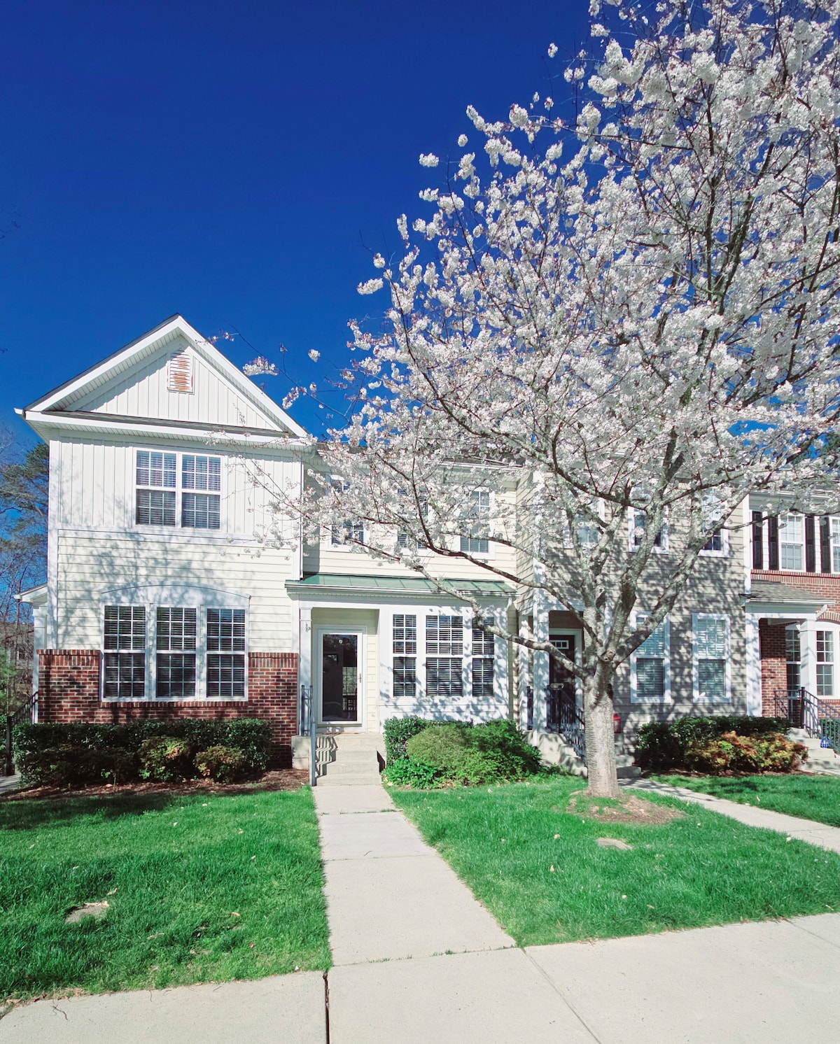 A charming building with light-colored siding and dark brick accents is framed by lush greenery. A flowering tree stands in front, enhancing the inviting appearance. A concrete pathway leads to the main entrance, which is flanked by multiple windows that allow natural light to fill the interior.
