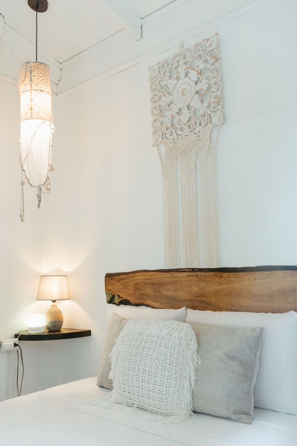 A cozy sleeping area features a spacious bed adorned with decorative pillows placed against a natural wood headboard. A macrame wall hanging adds texture above the bed. Soft lighting is provided by a pendant lamp and a bedside lamp, creating a peaceful ambiance.