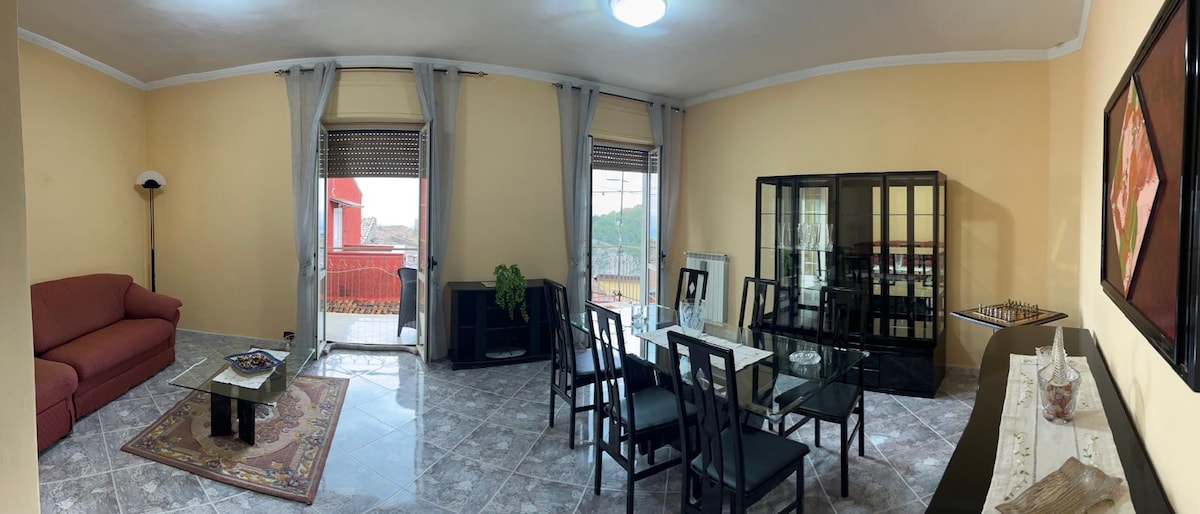 A spacious living area features a red sofa and a glass dining table surrounded by six black chairs. Natural light enters through multiple windows, with scenic views visible. A decorative area rug is placed in the center, complementing the neutral wall color.