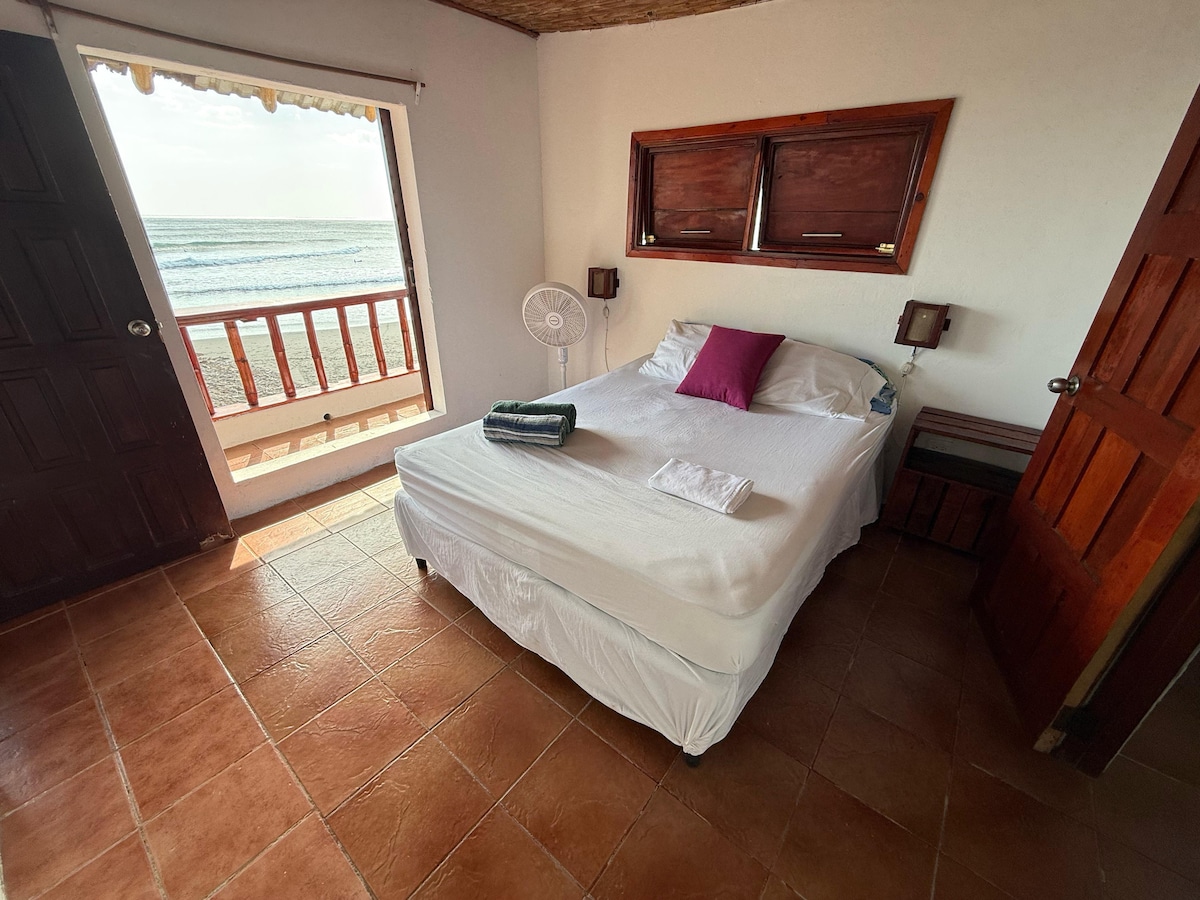 A cozy suite features a queen bed dressed in crisp linens, with a pillow accented in a warm hue. A private balcony overlooks the ocean, while a standing fan provides airflow. Two wooden doors offer access to the interior and sea view.