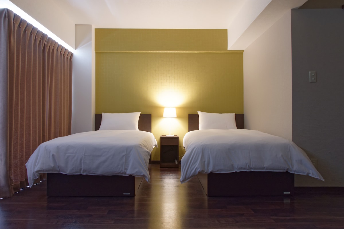 A well-lit room features two spacious beds adorned with crisp white linens. A nightstand with a lamp stands between the beds, while warm golden wall accents enhance the space. Light filters softly through sheer curtains, creating a calm ambiance.