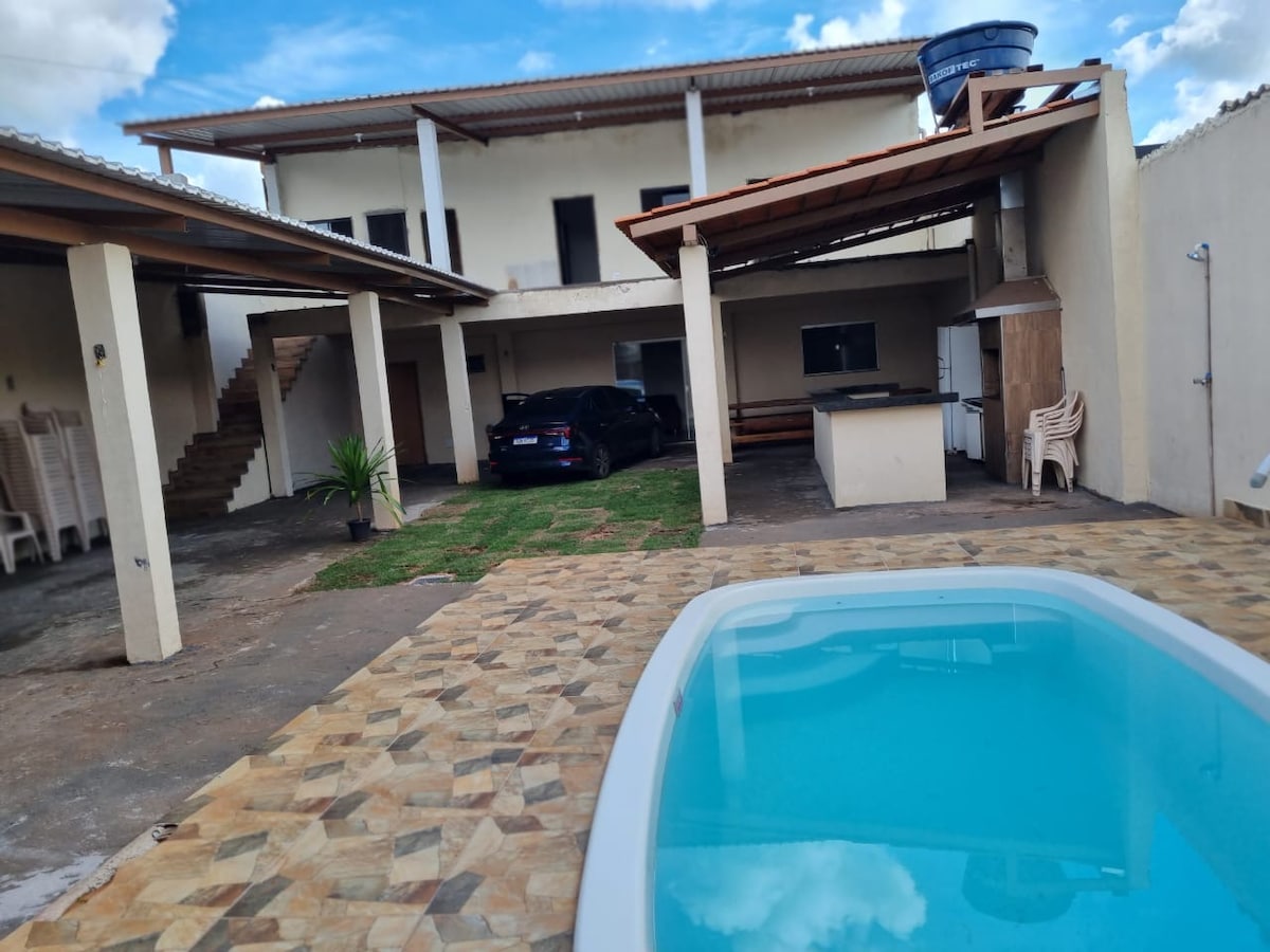An outdoor area featuring a clear swimming pool surrounded by patterned stone tiles. The backyard includes a car parked under a covered area, with a staircase visible leading to an upper level. Adjacent to the pool, lounging chairs are positioned for relaxation.