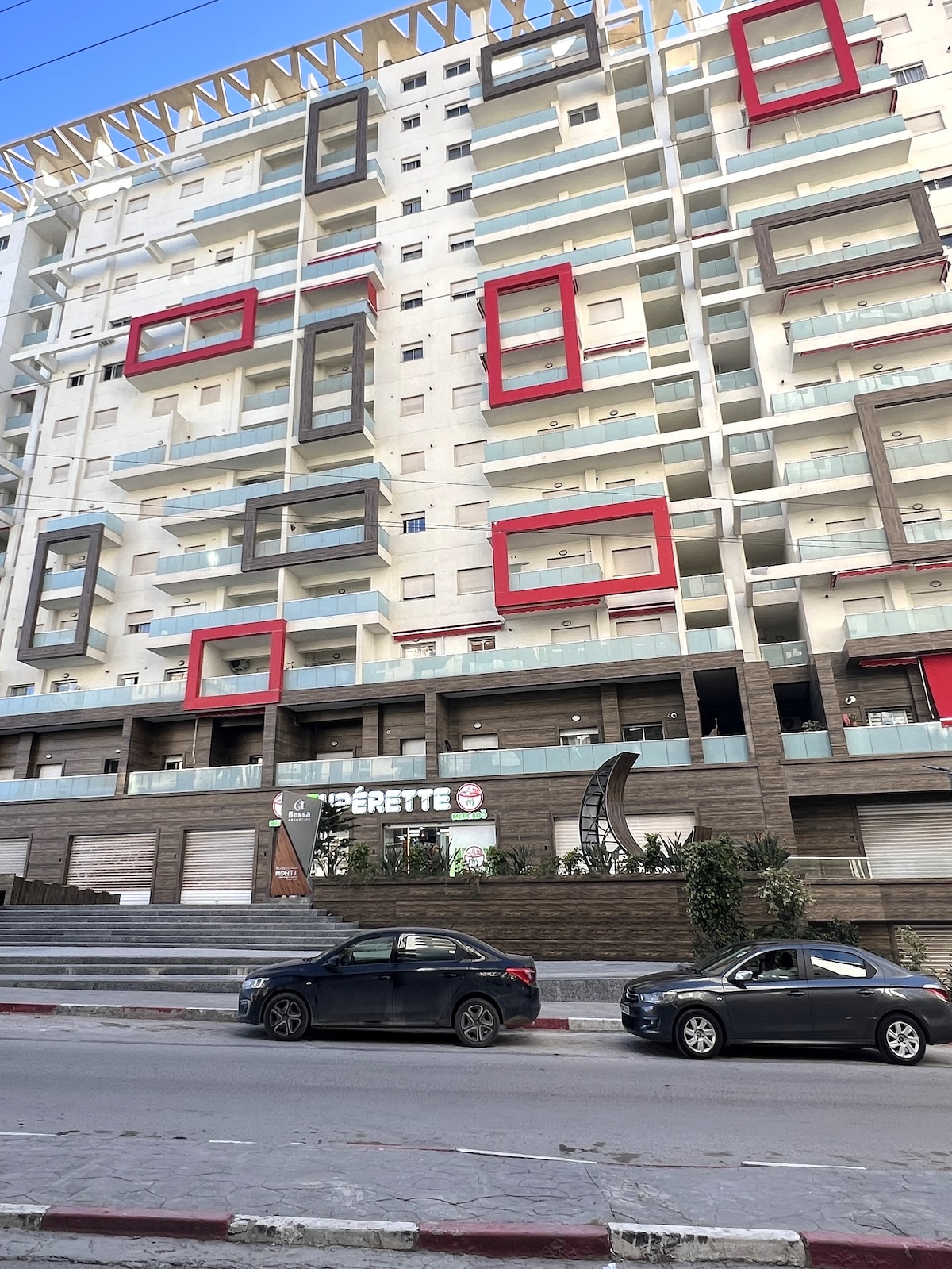 The contemporary building is showcased with a unique facade featuring a mix of balconies and large windows, framed by bold rectangular outlines in red and black. A smooth driveway is visible in front, with parked vehicles along the street.
