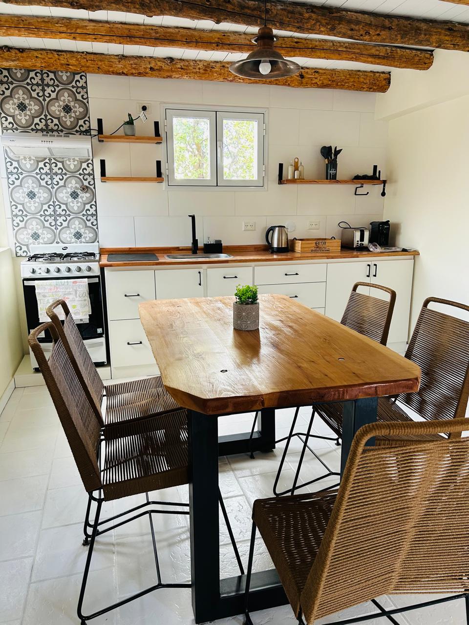 A modern kitchen area is highlighted by a wooden dining table surrounded by four metal-framed chairs. White cabinetry and a patterned backsplash are visible behind the stovetop. Natural light filters through a window, enhancing the clean, inviting space.