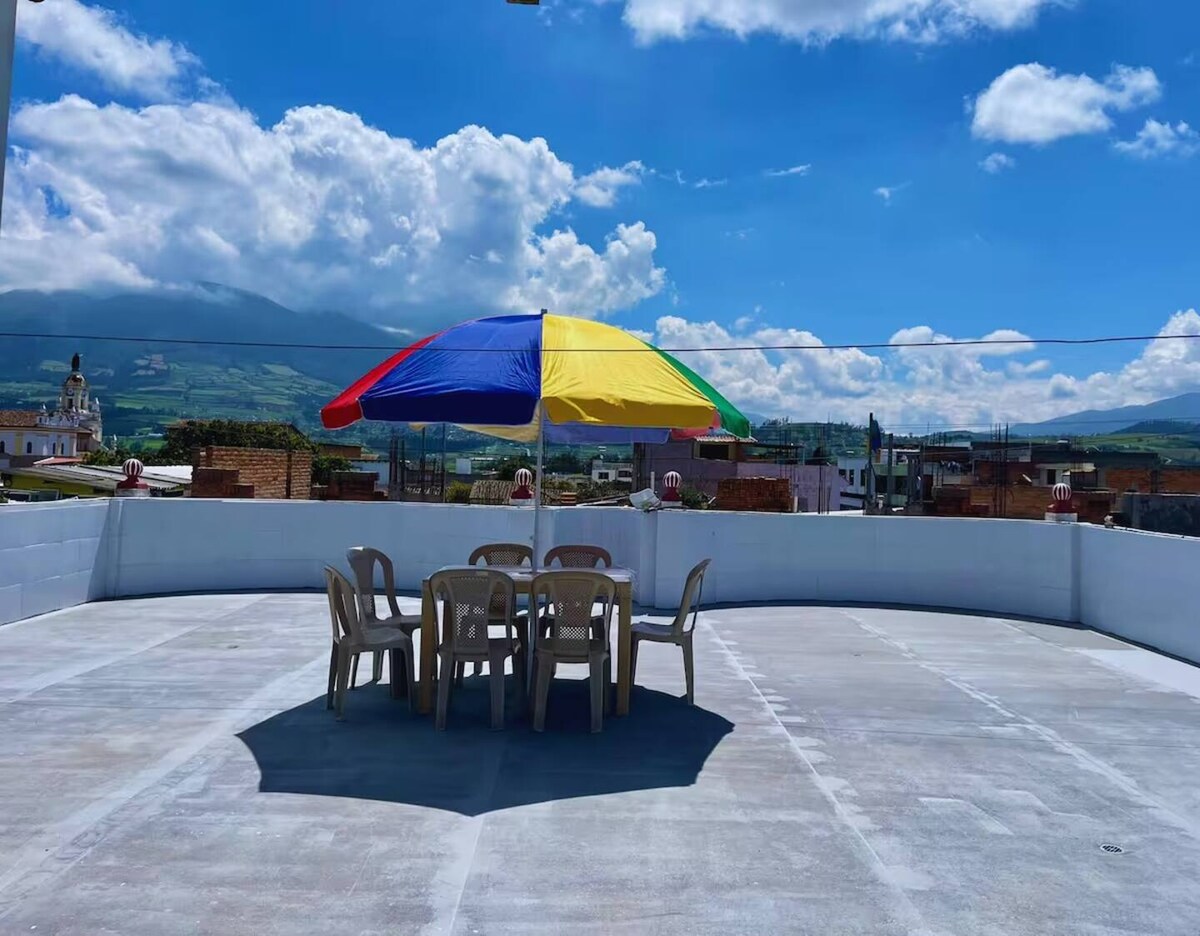 A spacious rooftop terrace is showcased with a colorful striped umbrella providing shade over a round table surrounded by several plastic chairs. The backdrop features clear skies and distant mountains, creating a serene setting for relaxation.
