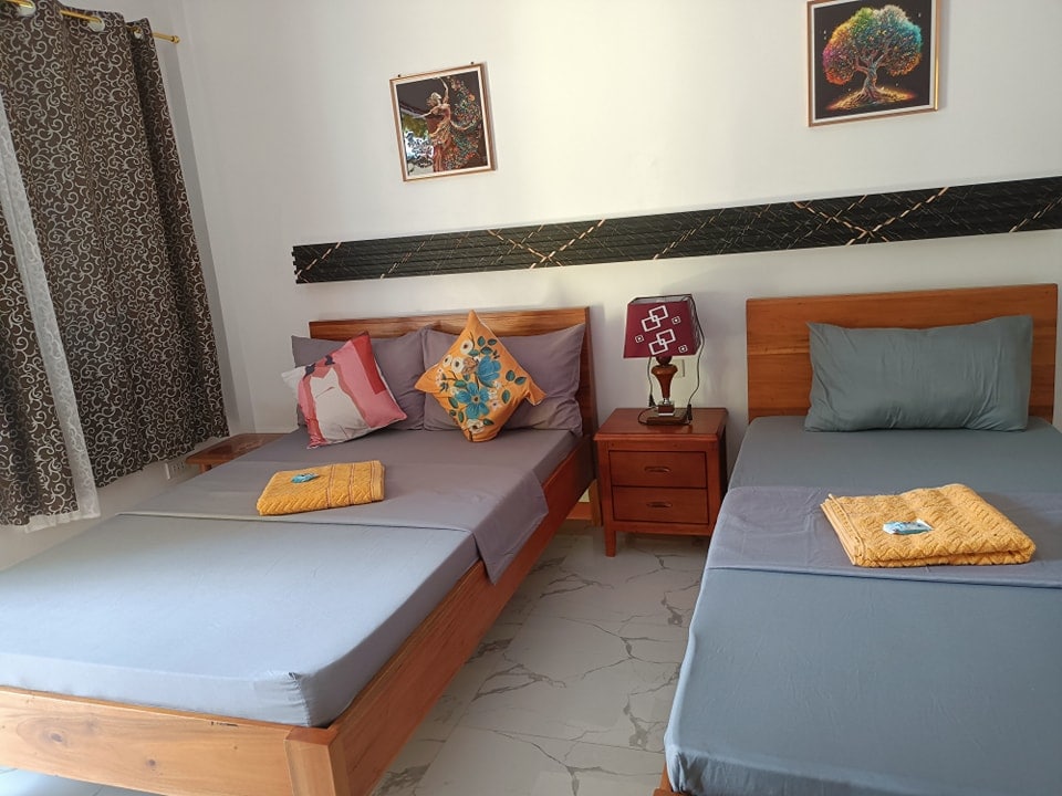 A cozy room is furnished with two spacious beds, each adorned with colorful pillows. Decorative wall art complements the light walls. A bedside table with a lamp is positioned next to one bed, while natural light filters through patterned curtains.