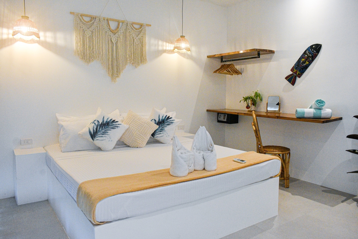 A serene bedroom features a spacious bed with decorative pillows and a light, neutral color scheme. Soft lighting is provided by hanging fixtures. A small workspace with a wooden table and chair is present, alongside a shelf displaying towels and decor, adding functionality to the space.