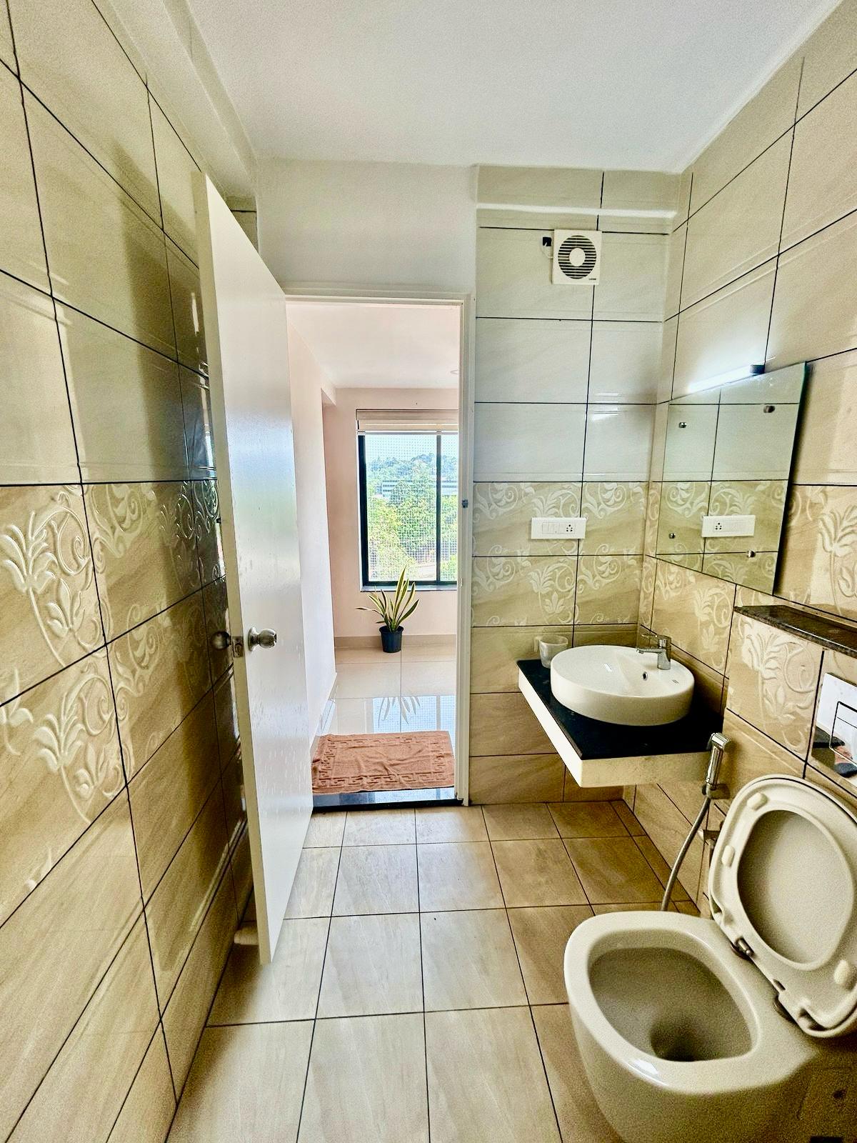 A well-appointed bathroom features beige tiles with floral patterns, a modern sink on a black countertop, and a toilet. A door leads to a well-lit space with a large window, allowing natural light to stream in, while a small plant adds a touch of greenery.