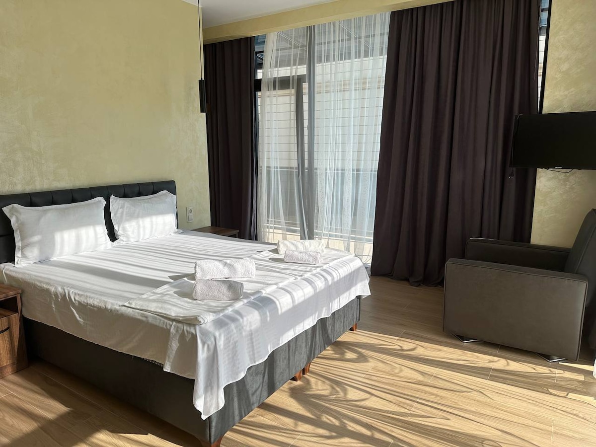 A bright sleeping area is highlighted by a spacious bed dressed in crisp white linens. Large paned windows allow natural light to fill the room, while dark curtains add contrast. A comfortable chair and a wall-mounted television complete the modern aesthetic.