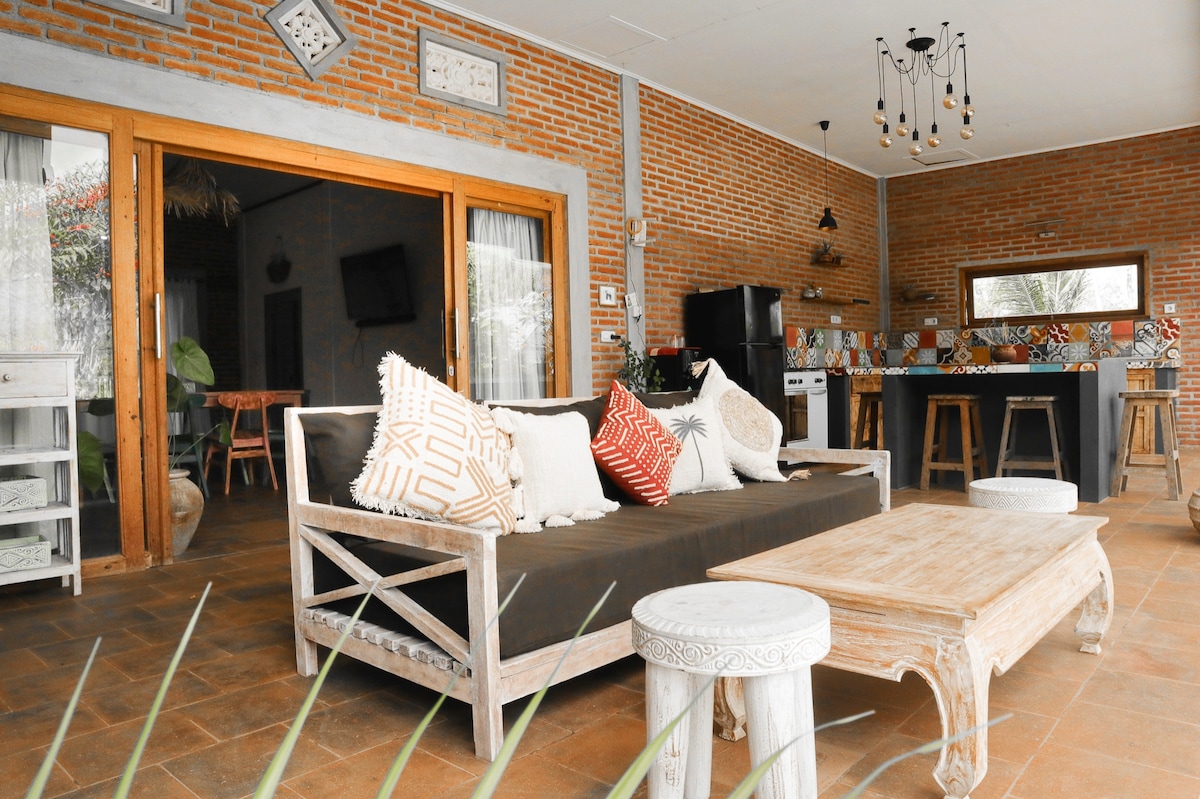 An open living area features a dark sofa adorned with decorative cushions, complemented by a light wooden coffee table. The space is framed by sliding glass doors, allowing natural light to flood in. A rustic kitchen area with wooden stools is visible in the background.
