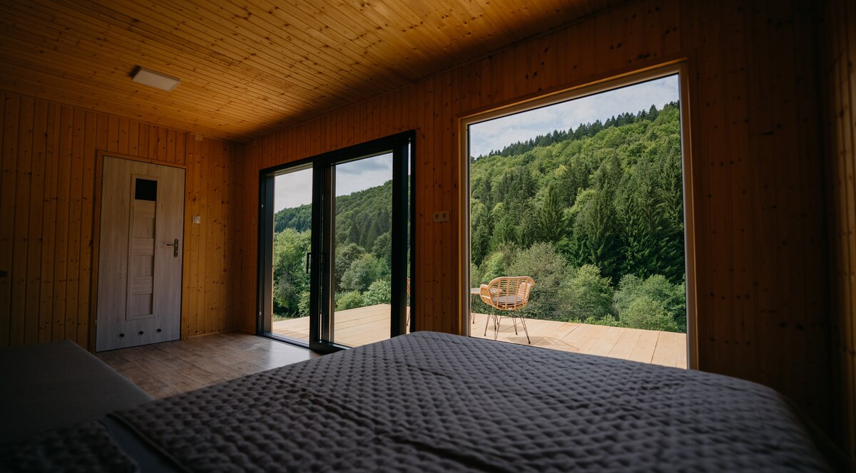 A room features wood-paneled walls and expansive glass doors that open to a deck. A spacious bed is positioned near the doors, offering views of the surrounding greenery. An accent chair sits on the deck, inviting relaxation amid nature.