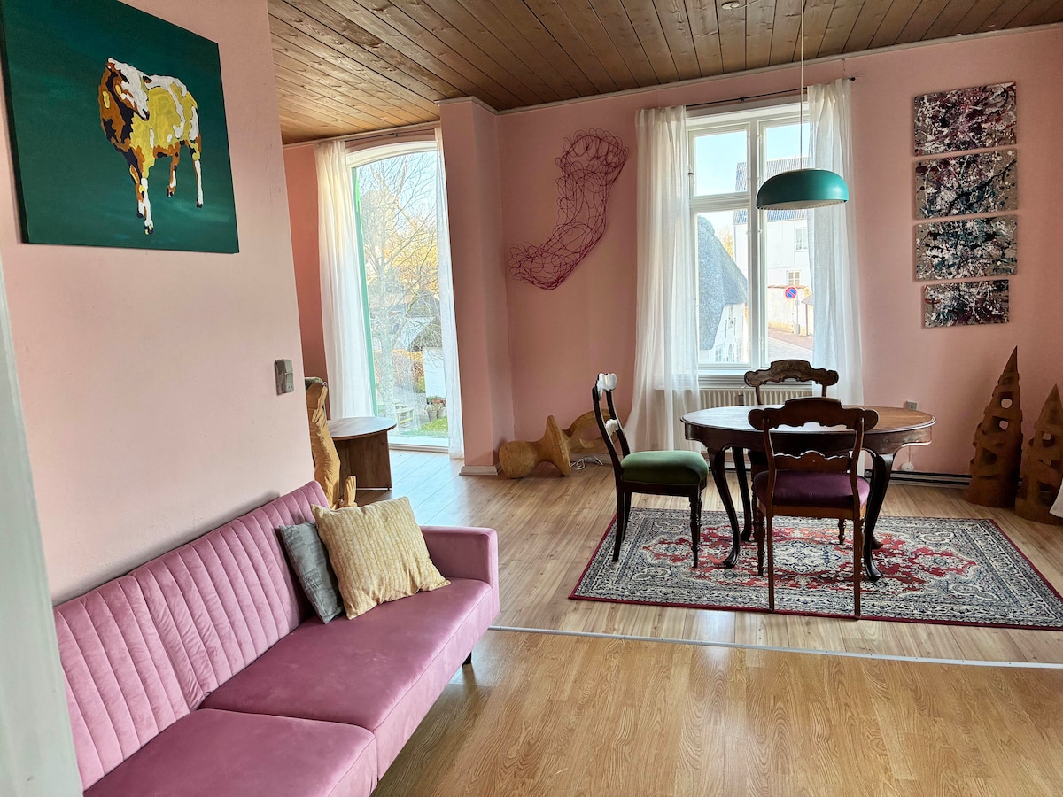 A living area showcases soft pink walls and a light wooden floor. A modern sofa in pastel pink is accompanied by colorful cushions. A round dining table is surrounded by four chairs, while artwork and decorative elements add character to the space. Natural light enters through large windows.