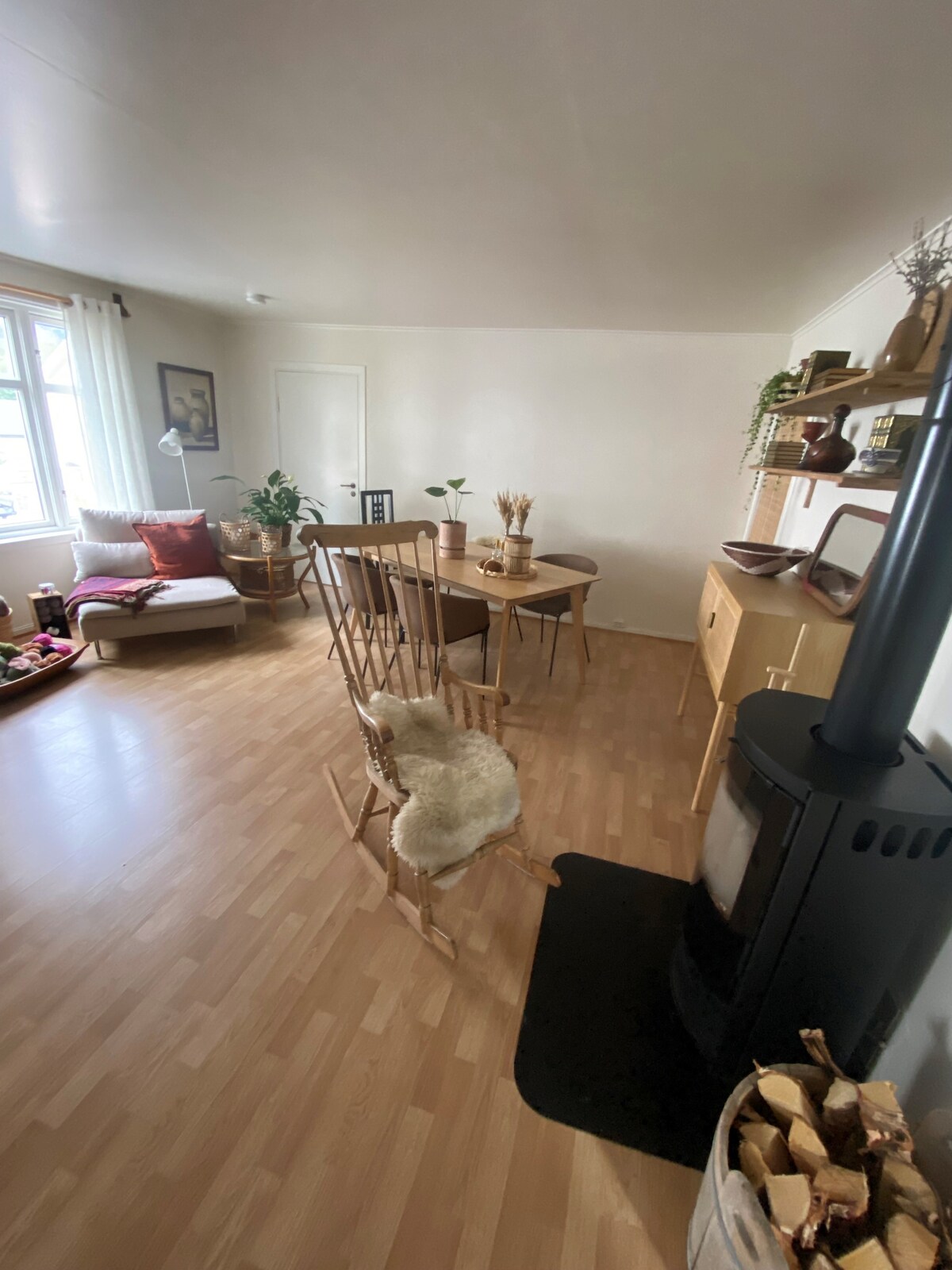 A cozy living area features a mix of seating options, including a sofa with cushions and a dining table surrounded by chairs. A wood-burning stove stands in the corner, with a stack of firewood nearby. Natural light enters through a window, illuminating the space.