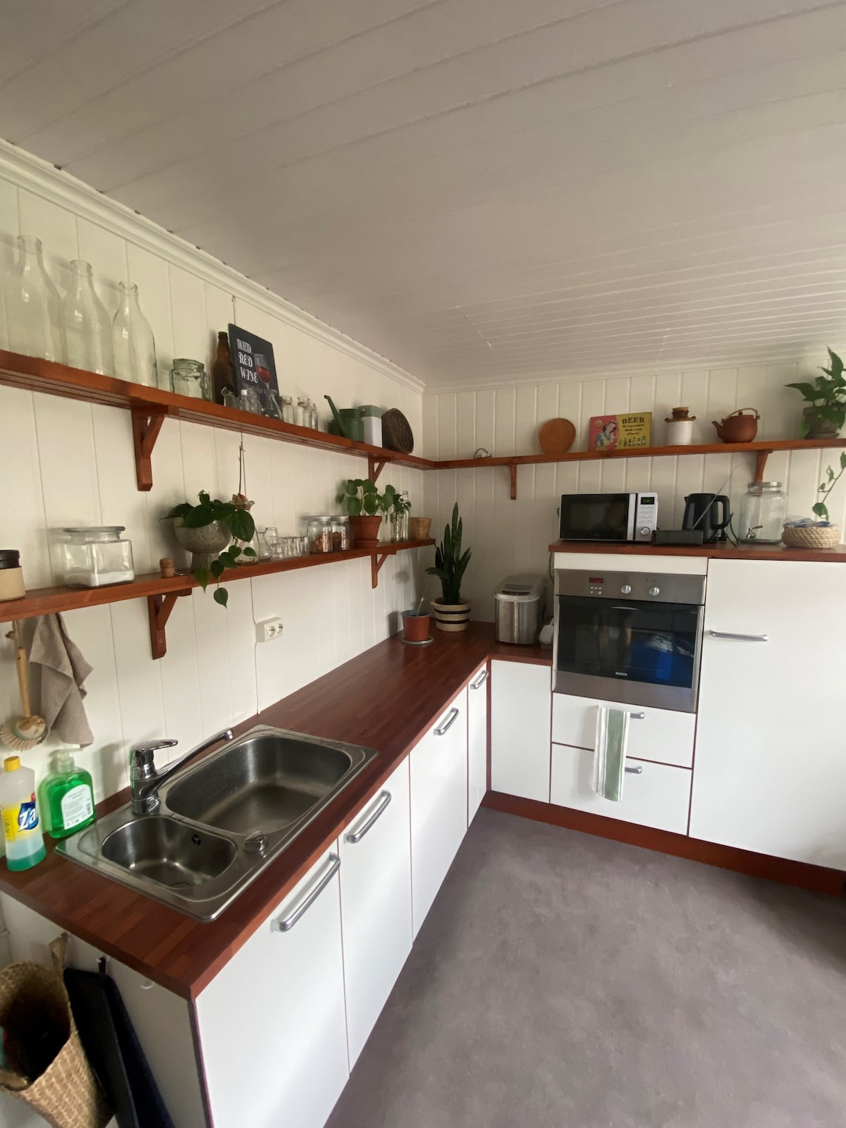 A well-organized kitchen features light-colored cabinetry and a wooden countertop. Shelves display an assortment of glassware and potted plants. Appliances include a microwave and a compact oven, while a double sink is positioned under the windows. Kitchen essentials are neatly arranged on the counter.