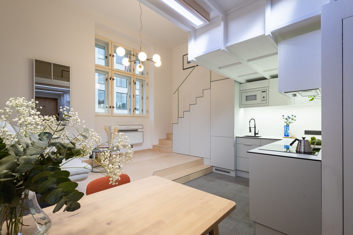 A modern kitchen area is seen with a minimalist design, featuring sleek cabinetry and a stainless-steel sink. A wooden dining table with simple chairs is positioned in the foreground. Light floods in through large windows, and decorative flowers add a touch of greenery to the space.