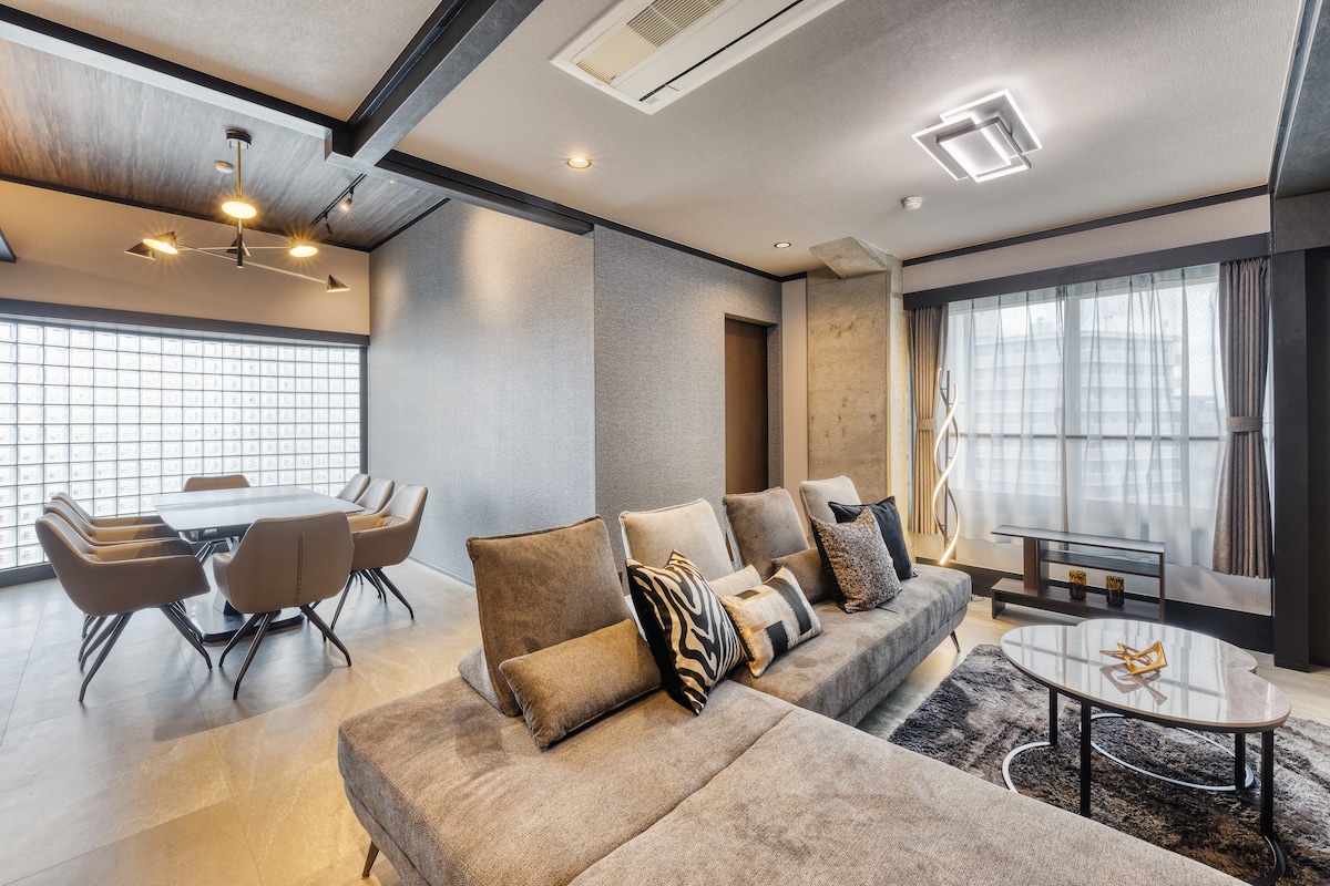 A spacious living area is furnished with a modern sectional sofa, complemented by decorative cushions. A coffee table is placed centrally, while a dining area features stylish chairs around a rectangular table. Natural light filters through large windows, creating an airy ambiance.