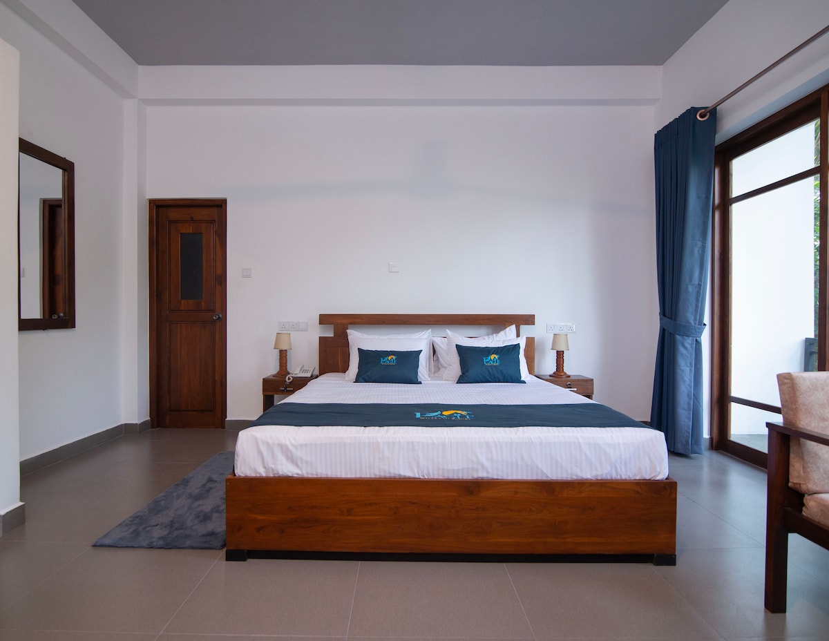 A spacious room features a king-sized bed with crisp linens and two bedside tables. Natural light floods the space through large windows adorned with blue curtains. A wooden mirror reflects the minimalistic decor, complemented by a comfortable chair placed beside the bed.
