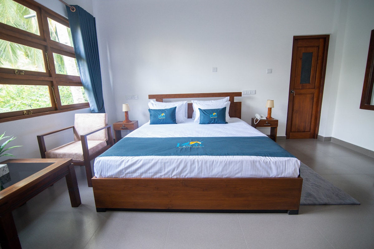 A spacious room features a large bed with crisp white linens and accent pillows in shades of blue. Natural light floods in through large windows adorned with dark curtains, while a comfortable armchair and a small side table enhance the inviting decor.