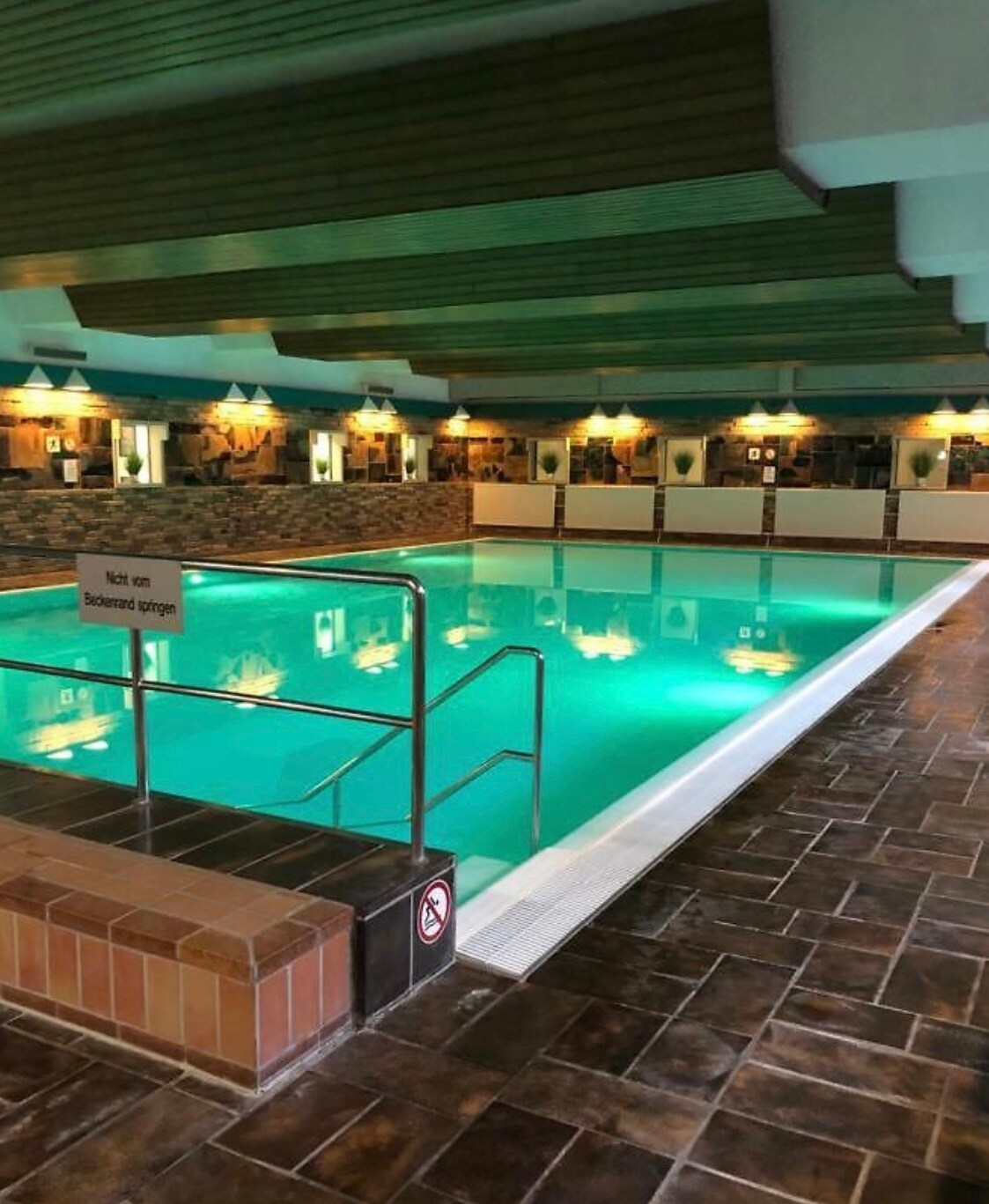 An indoor pool is framed by stone walls and wooden ceilings. The clear turquoise water reflects soft lighting from the surrounding fixtures. Handrails provide accessibility, and safety signage is visible nearby, ensuring a safe swimming environment.