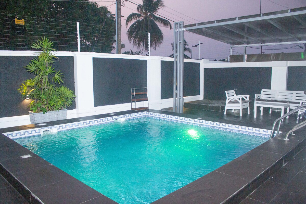 A private swimming pool is surrounded by dark tiled flooring and a white boundary wall. Green lighting illuminates the water, creating a calm ambiance. Nearby, a set of white outdoor chairs and a small table are positioned against a backdrop of palm trees.