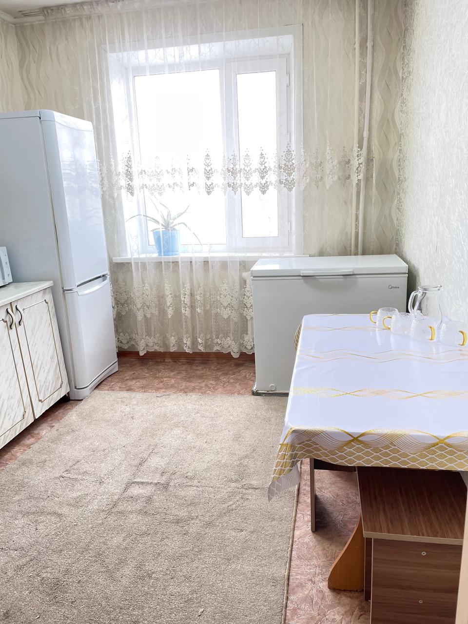 The kitchen features a white refrigerator and a spacious chest freezer. A window dressed with light lace curtains allows natural light to fill the room. A simple table with a decorative tablecloth sits beside a small wooden kitchen desk, enhancing the functional layout.