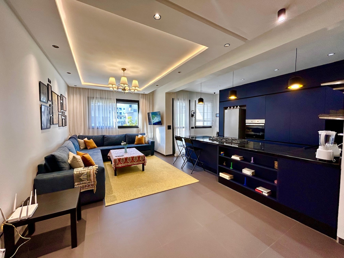 A bright and modern living space features a large sectional sofa draped with decorative pillows. A coffee table centers the room, complemented by a textured area rug. The open kitchen, equipped with dark cabinetry and sleek appliances, is visible in the background.