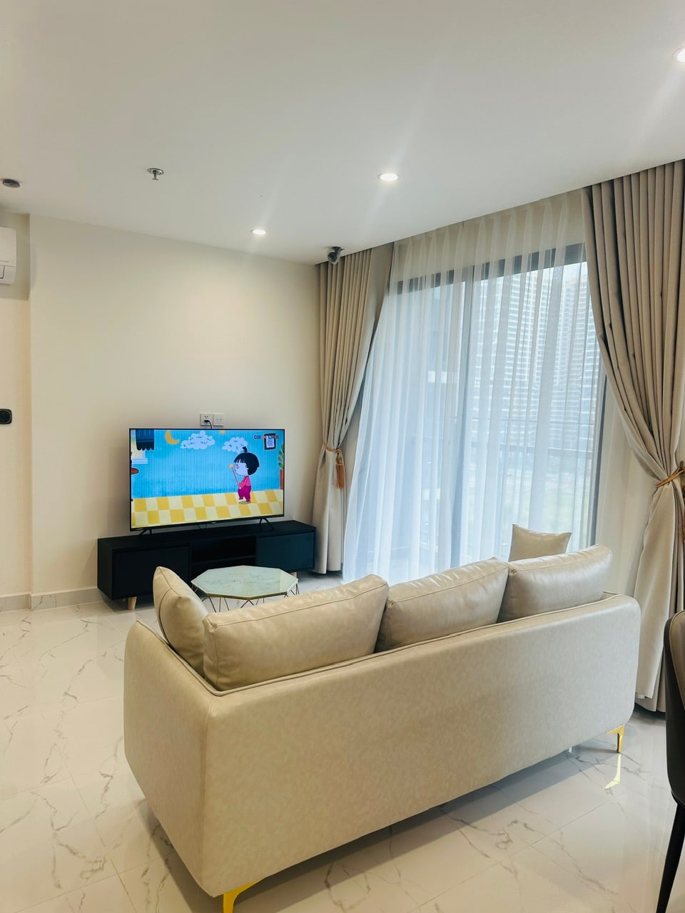 A light-filled living area features a comfortable sofa facing a wall-mounted television. Sheer curtains frame large windows, allowing soft natural light to illuminate the space. A small round table sits in front of the sofa, enhancing the room's welcoming atmosphere.