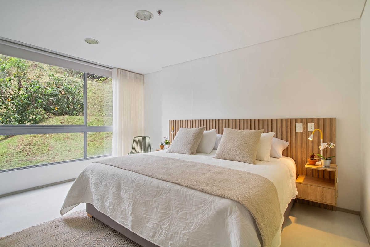 A spacious bedroom features a large bed with neutral-toned bedding and several cushions, complemented by a wooden headboard. Natural light pours in through a large window, offering a view of greenery outside. A side table with a lamp adds functionality to the room.
