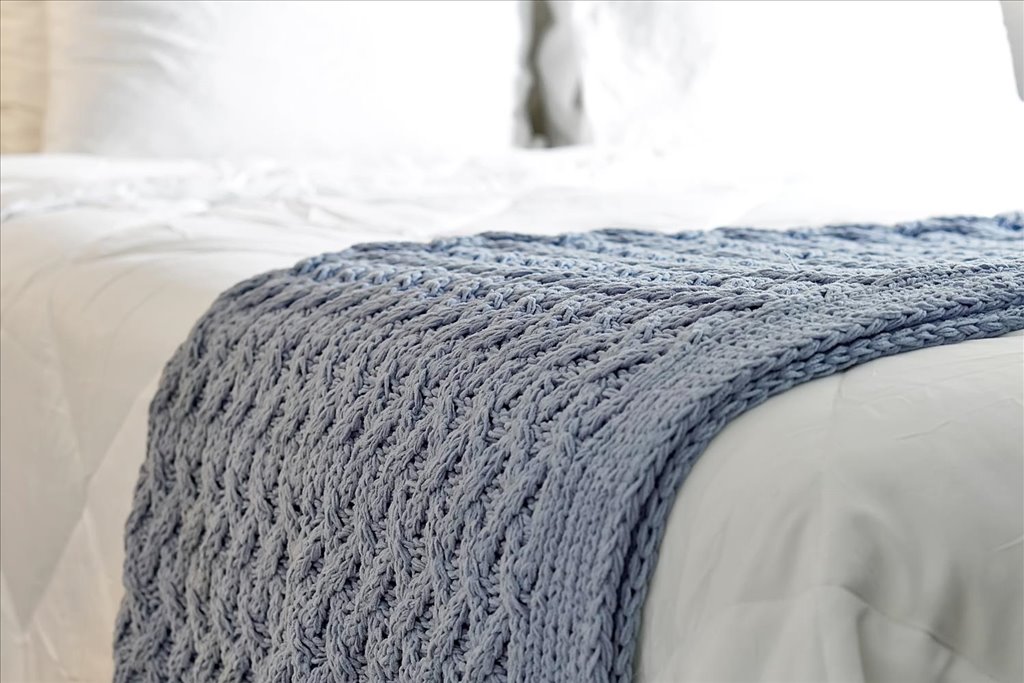 A textured light blue knitted throw blanket is draped across a neatly made bed. The bed is dressed in soft white linens, creating a calm and inviting look. Light illuminates the space, emphasizing the blanket's intricate design.