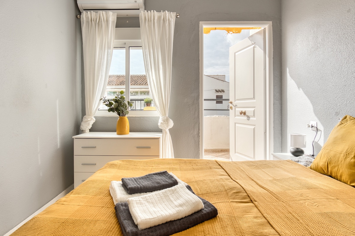 A serene bedroom features a double bed with a textured mustard-yellow bedspread, complemented by two neatly folded towels. Natural light filters through sheer curtains, illuminating a white dresser and a potted plant. A door leads to the balcony, enhancing the tranquil ambiance.