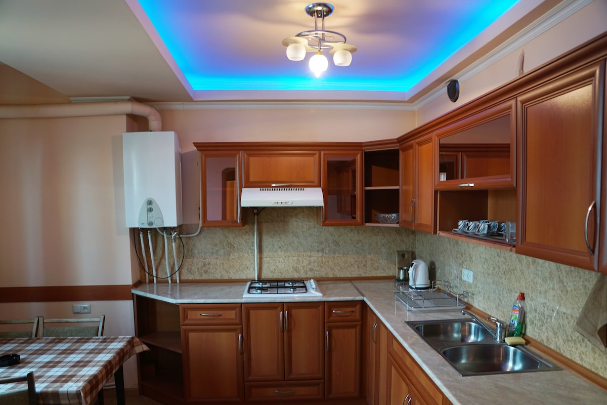A well-equipped kitchen features wooden cabinets and a light marble countertop. Modern appliances, including a stovetop and kettle, are visible. A dining table with a checkered tablecloth is positioned to one side, while ambient lighting casts a soft glow over the space.