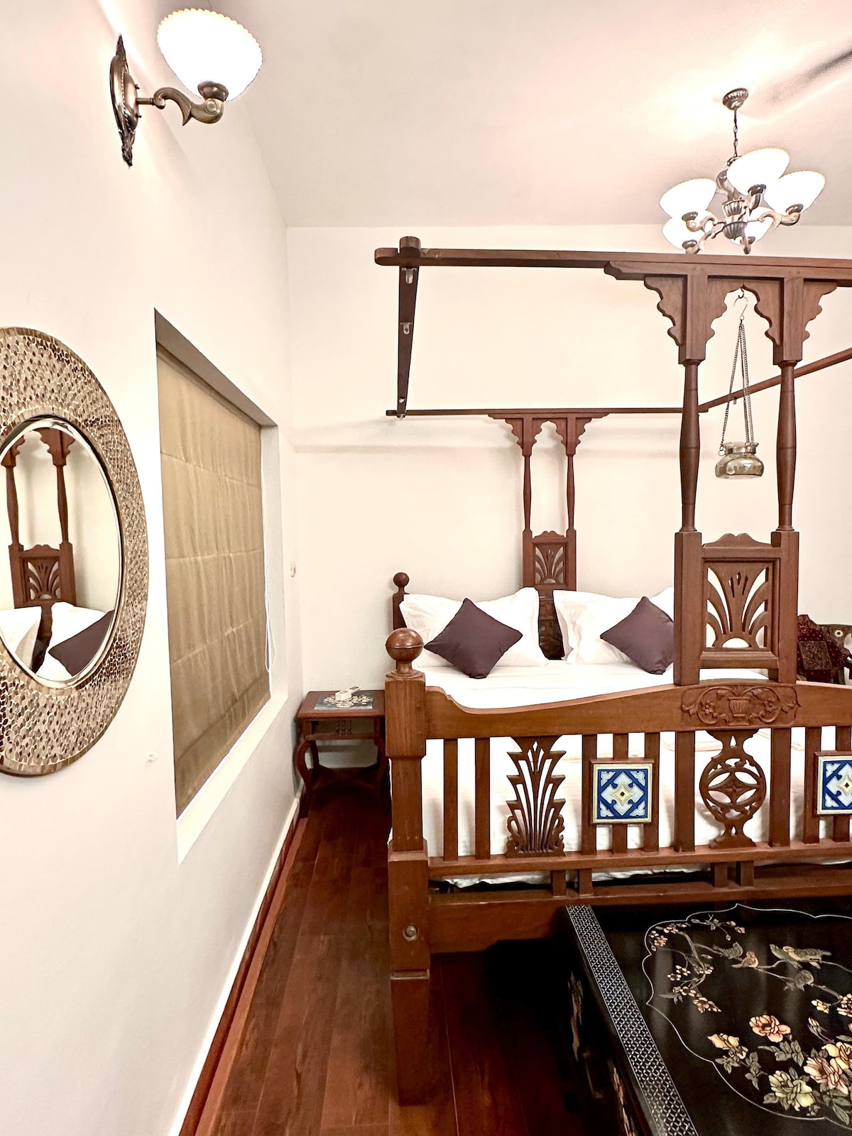 A wooden four-poster bed is adorned with decorative elements and complemented by soft pillows. A round mirror hangs above a bedside table, while a lantern adds a warm glow. Rich wooden flooring enhances the serene ambiance in this cozy private bedroom.
