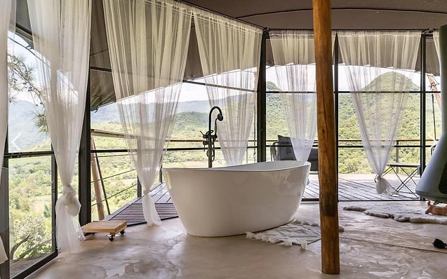 A freestanding bathtub is positioned on a terrace surrounded by sheer curtains, offering an unobstructed view of the mountains. The smooth concrete floor is complemented by soft rugs, while wooden posts support the structure, creating an open and airy atmosphere.