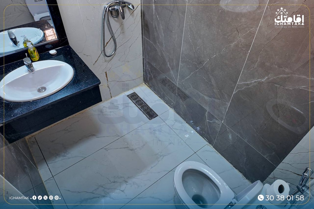 A modern bathroom features sleek marble walls and floor tiles. A clean sink is positioned above a minimalist countertop, while a large mirror reflects the space. A showerhead is mounted against the wall, and a toilet is positioned beside a drainage grate on the floor.