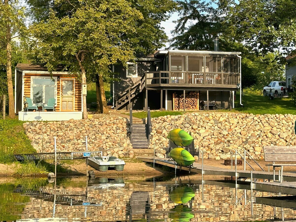 A lakeside cabin and modern structure are depicted, surrounded by greenery. A stone path leads to a wooden deck, where stacked firewood is visible. Two kayaks sit on the shore, reflecting in still water, enhancing the serene lakeside environment.