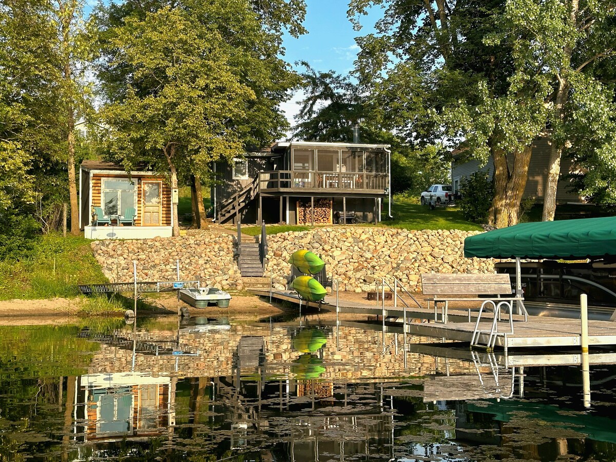 A lakeside retreat is visible, featuring a multi-level structure surrounded by lush trees. A stacked stone wall leads down to a dock equipped with seating. Reflection on the water creates a serene atmosphere, with a small boat and green lily pads gently resting nearby.